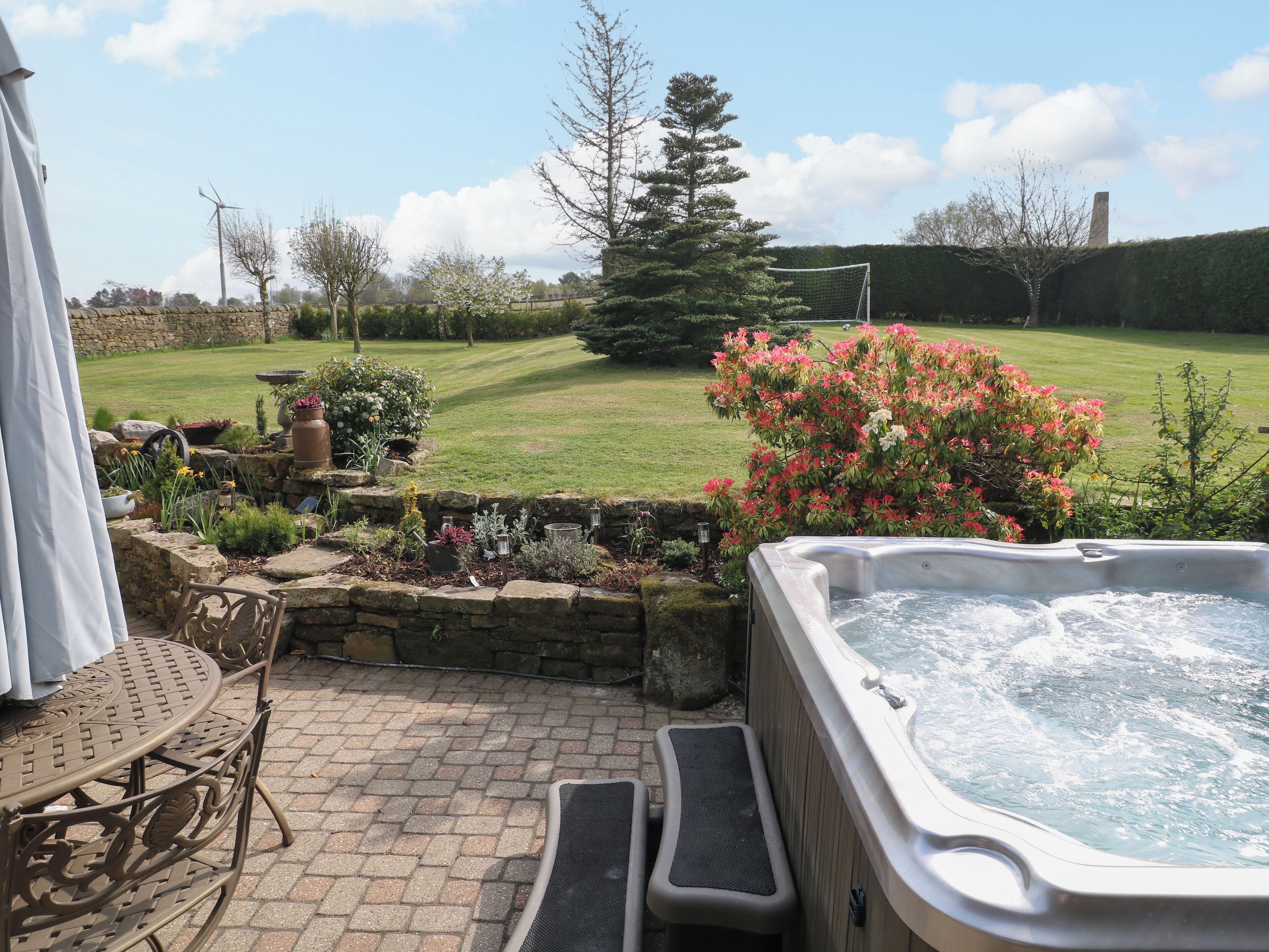A hot tub in a garden with a stone wall, a chair, and a lawn.