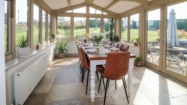A conservatory with a dining table set for six, surrounded by large windows offering a view of a garden. - Cherry Tree Farm (Chesterfield)
