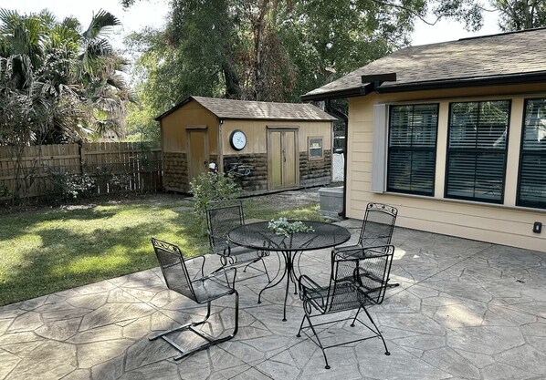 Une terrasse avec une table ronde et des chaises, un petit abri de jardin et une maison dotée de grandes fenêtres.