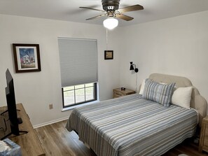 Une chambre à coucher comprenant un lit, un ventilateur de plafond, une fenêtre avec des stores, une table de chevet et une télévision.