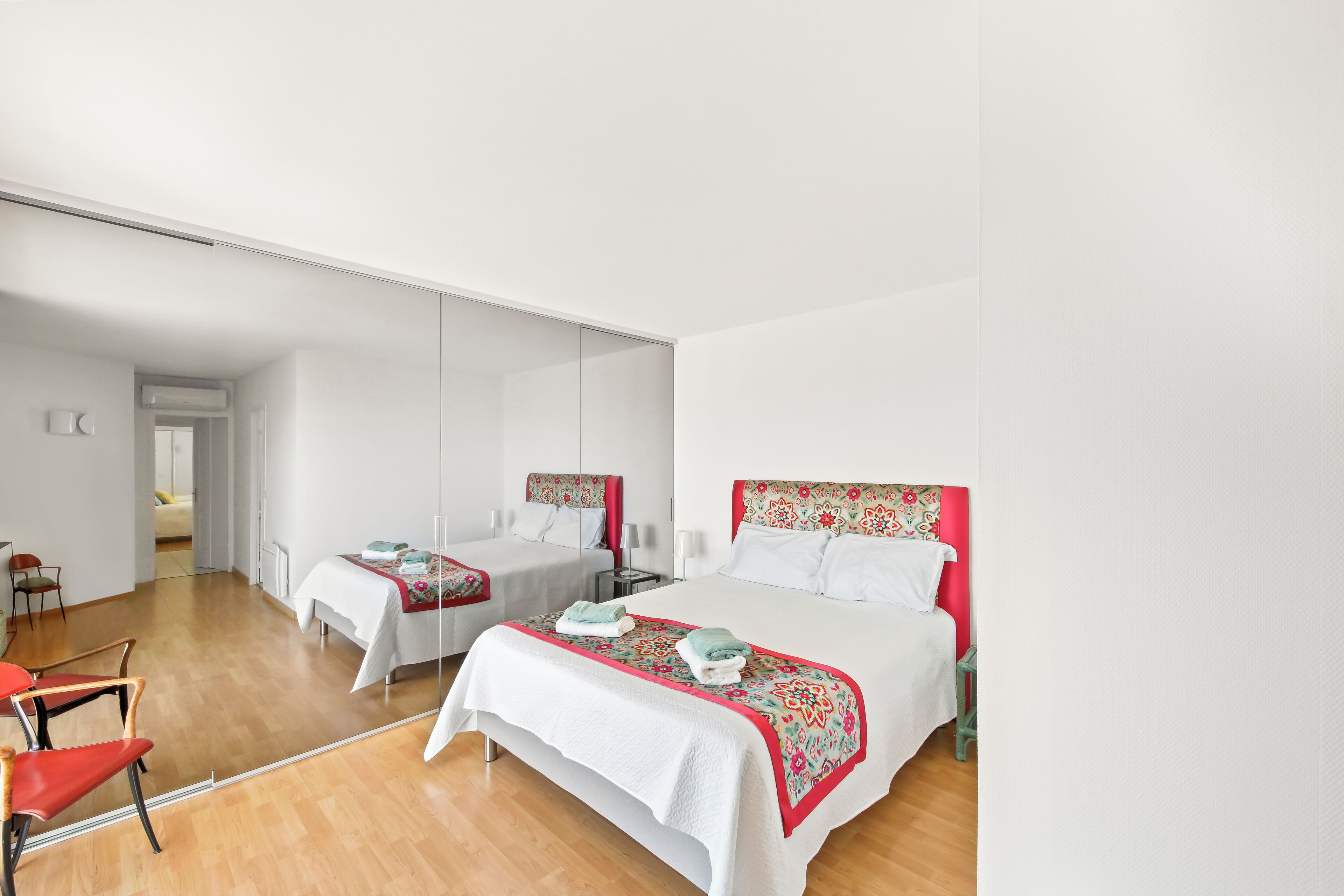 Ein modernes Hotelzimmer mit zwei Betten, einem großen Spiegel, Holzboden und einem rot-weißen gemusterten Kopfteil.