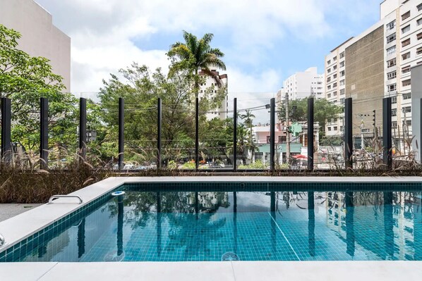 A swimming pool with a tiled deck, surrounded by a fence and trees, with buildings in the background. - Cardosolf in S o Paulo (São Paulo)