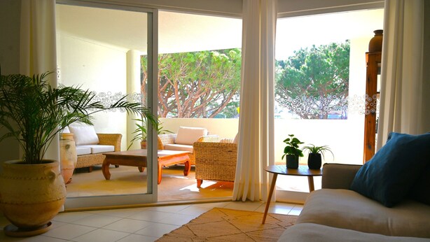 A living room with a sofa, a coffee table, and a large potted plant. There is a sliding glass door leading to an outdoor area with wicker furniture and a view of trees. - Jardins do Golfe at Vale do Lobo (Loulé)