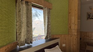 A bedroom with a wooden headboard, a bed with white pillows, a window with floral curtains, and a view of a mountain. - Nature Heaven Homestay (Shimla)