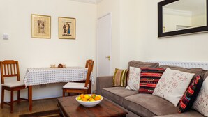 A living room with a sofa, a dining table with chairs, a bowl of fruit, and two framed pictures on the wall.