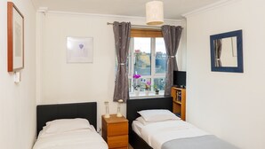 A bedroom with two beds, a bedside table, a lamp, a window with curtains, a mirror, and a framed picture on the wall.