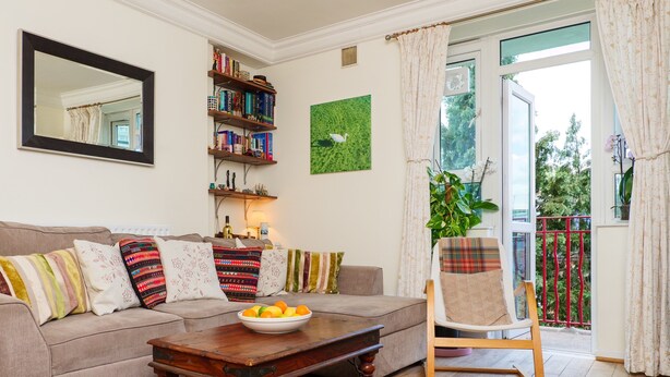 A living room with a sofa, a wooden coffee table with a bowl of fruit, a bookshelf, a painting, and a view of a balcony with a plant.