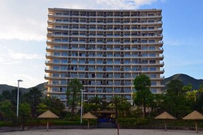 Un hôtel à plusieurs étages, doté de balcons, situé près d’une plage bordée de palmiers et parsemée de parasols.