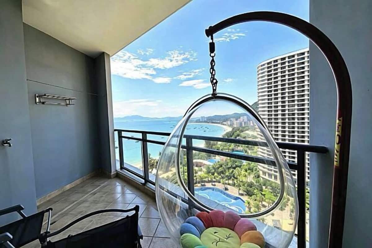 A balcony with a hanging chair, a view of a beach, and a pool.