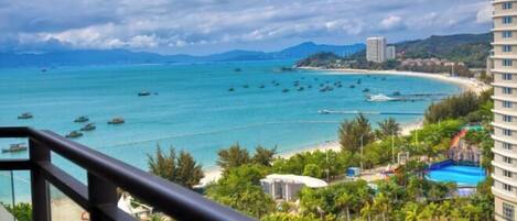 A coastal view from a balcony with a beach, boats, and a resort area including a pool and a water slide.