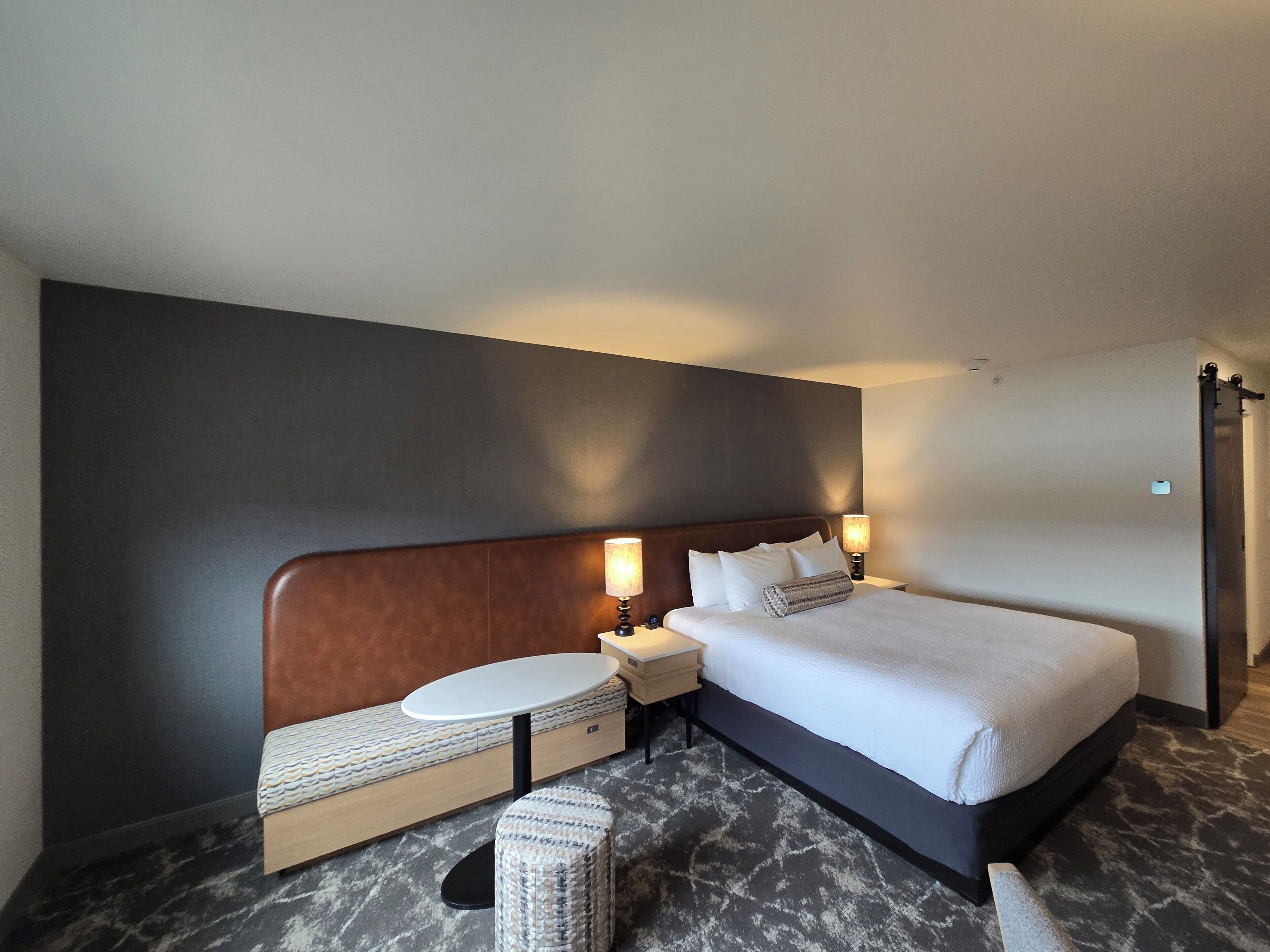A modern hotel room with a large bed, bedside tables, a round table, and a bench.