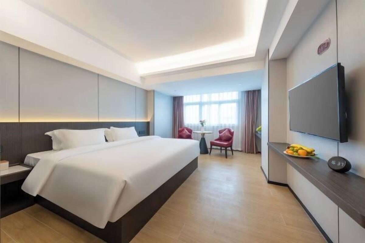A hotel room with a large bed, a TV, a desk with a chair, and a small table with fruit.