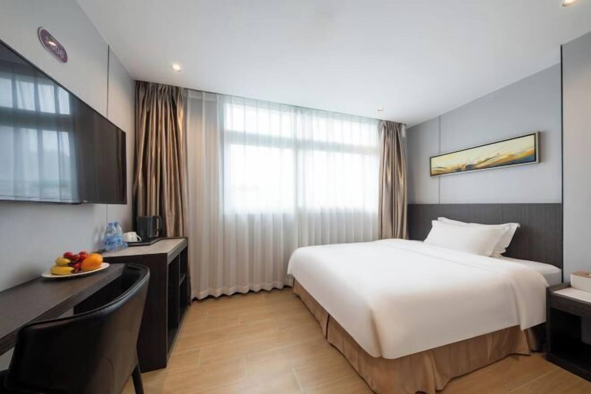 A hotel room with a bed, a desk with a chair, a TV, a painting on the wall, and a window with curtains.