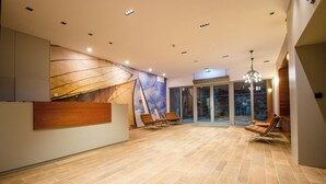 A spacious lobby with a wooden reception desk, two lounge chairs, a large mural, and a view through glass doors. - Waterlane Island Deluxe 5B by GrandApart (Gdansk)