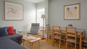 Een woonkamer met een grijze bank, een houten eettafel met stoelen, een klein rond tafeltje met een tijdschrift en twee ingelijste kunstwerken aan de muur.
