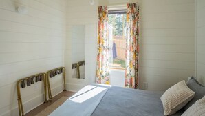 A bedroom with a bed, a window with floral curtains, a mirror, and a bedside table.