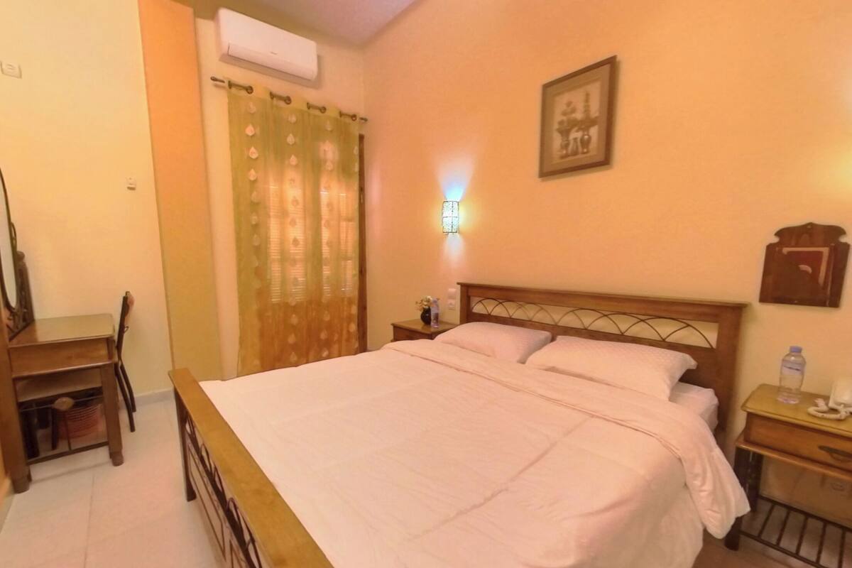 A bedroom with a bed, a desk, a chair, a nightstand, and a wall-mounted lamp.