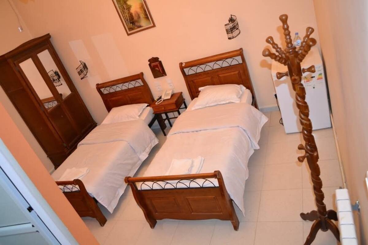 A room with two wooden beds, a small table with a telephone, a coat rack, and a refrigerator.