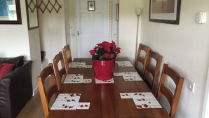 A dining area with a wooden table set for four, surrounded by wooden chairs. A red pot with red flowers is placed in the center of the table. - 3 bed Detached Sound Healing Retreat Bungalow (Cardigan)