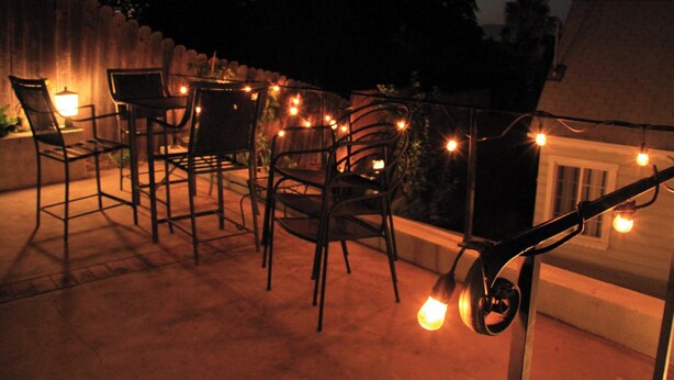 Une terrasse extérieure avec des chaises et des tables en métal, des guirlandes lumineuses et une vue sur une maison en arrière-plan.
