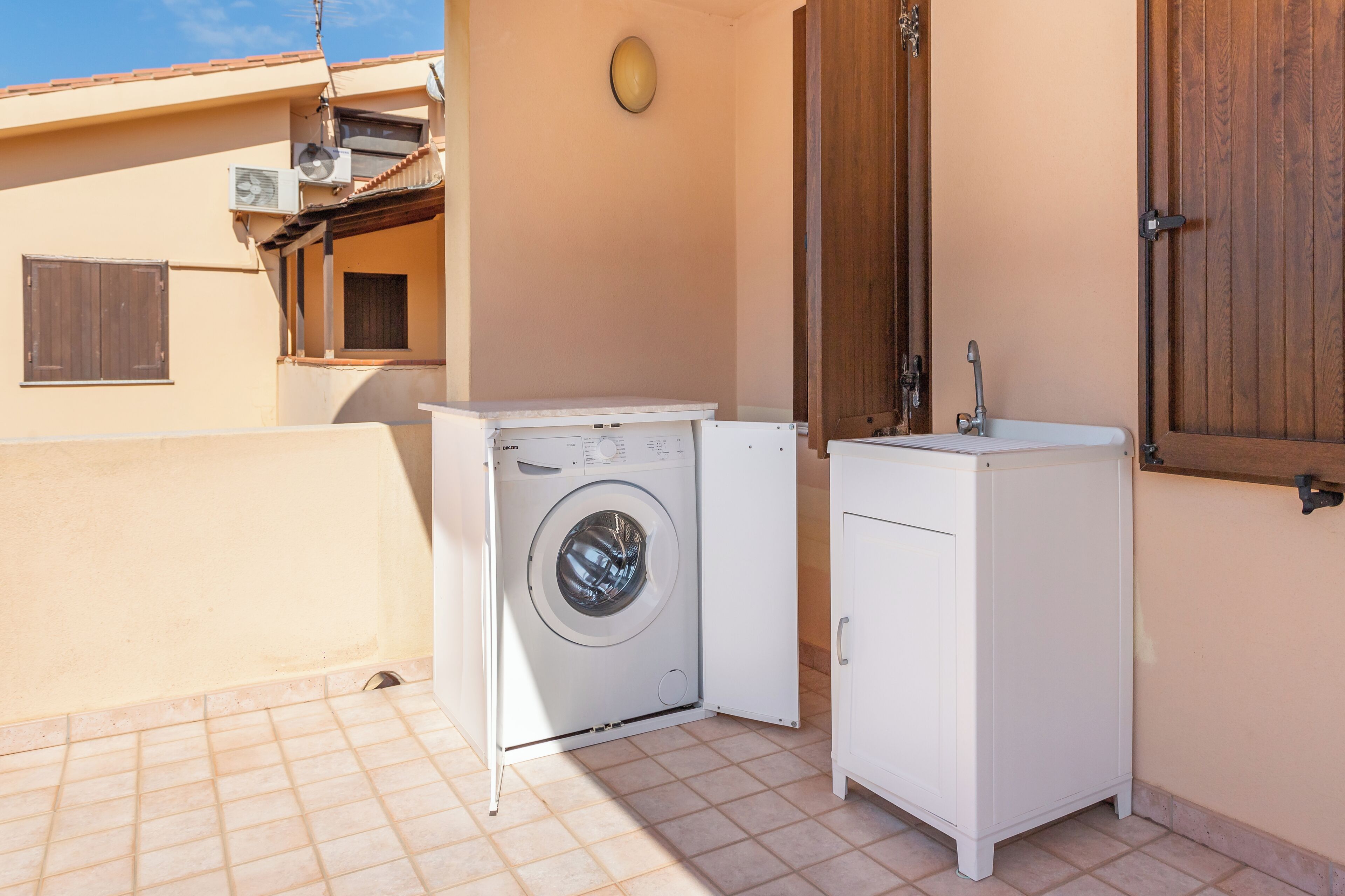 A compact laundry area with a washing machine and a small sink, adjacent to a building with a tiled floor and a wooden door.