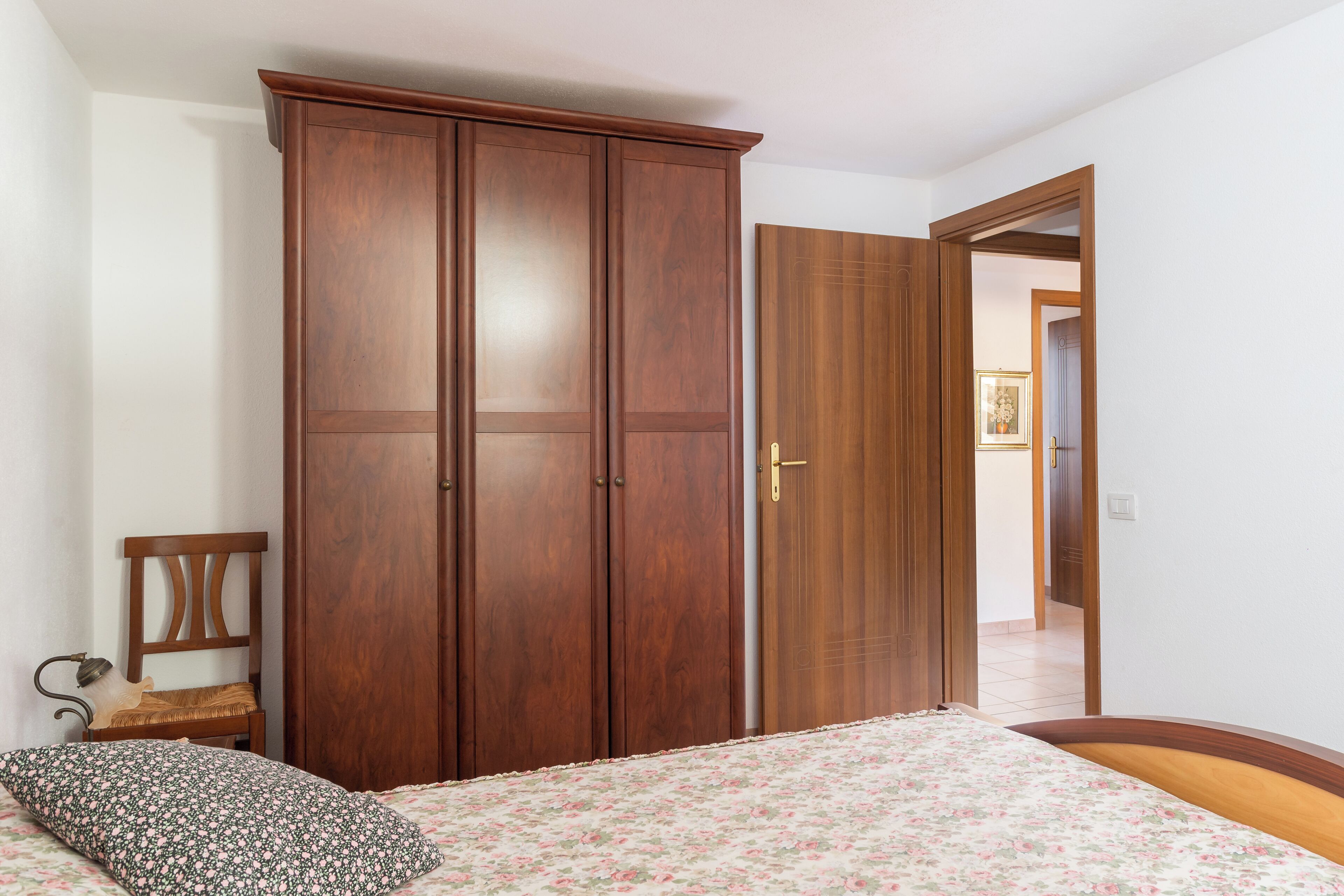 Une chambre à coucher avec une armoire en bois, un lit recouvert d’une courtepointe à motifs floraux et une chaise avec un coussin.