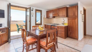 Une cuisine avec des meubles en bois, une table à manger avec des chaises, un réfrigérateur et une vue sur l’extérieur.