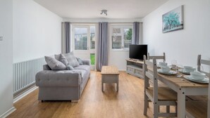 A modern living room with a grey sofa, a wooden coffee table, and a dining area with a wooden table and chairs. - Gatwick Layover: Modern 1BR Free Parking & Garden (Crawley)