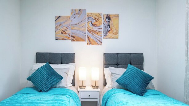 A twin room with two beds, each with a headboard, a nightstand, and a lamp. There are three wall-mounted artworks above the beds. - Gatwick Layover: Modern 1BR Free Parking & Garden (Crawley)