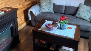 En mysig stuga med träpanel på väggarna, en grå soffa, ett soffbord med en korg och blommor, och en inramad tavla på väggen.