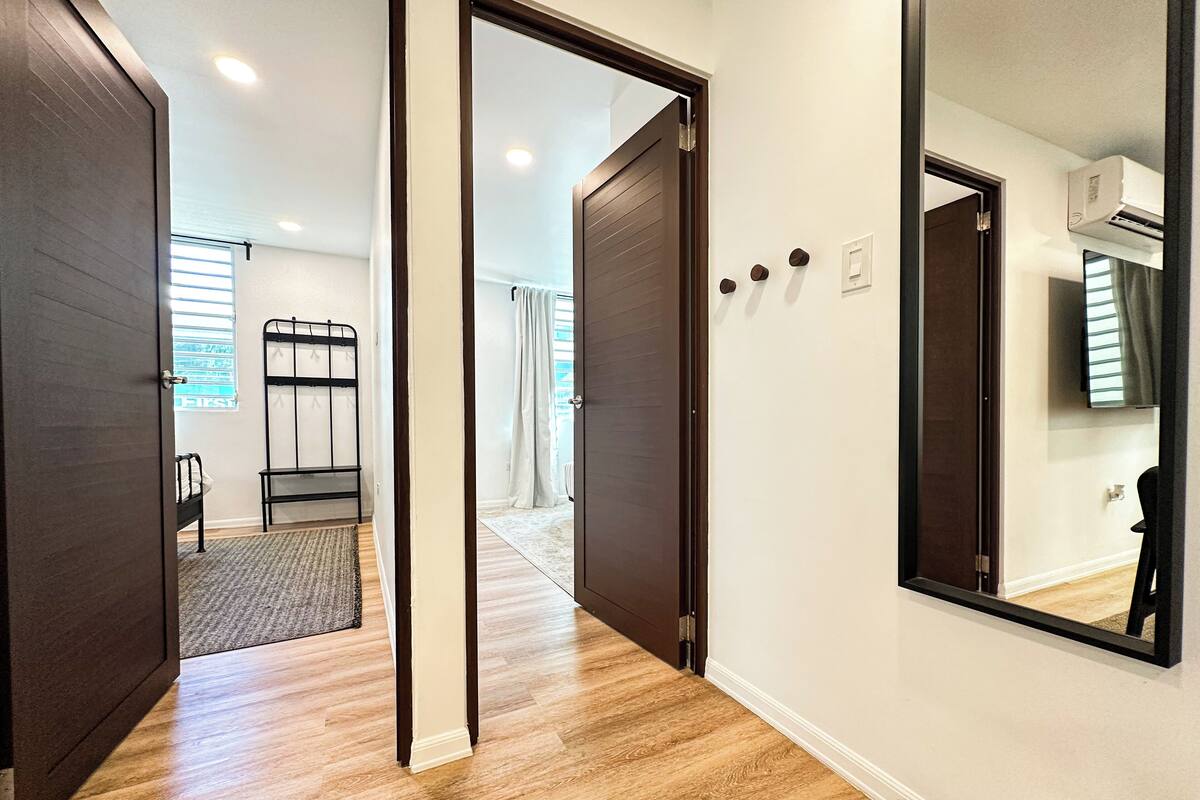 A hallway with wooden flooring, a bed, a mirror, and a door leading to a bathroom.