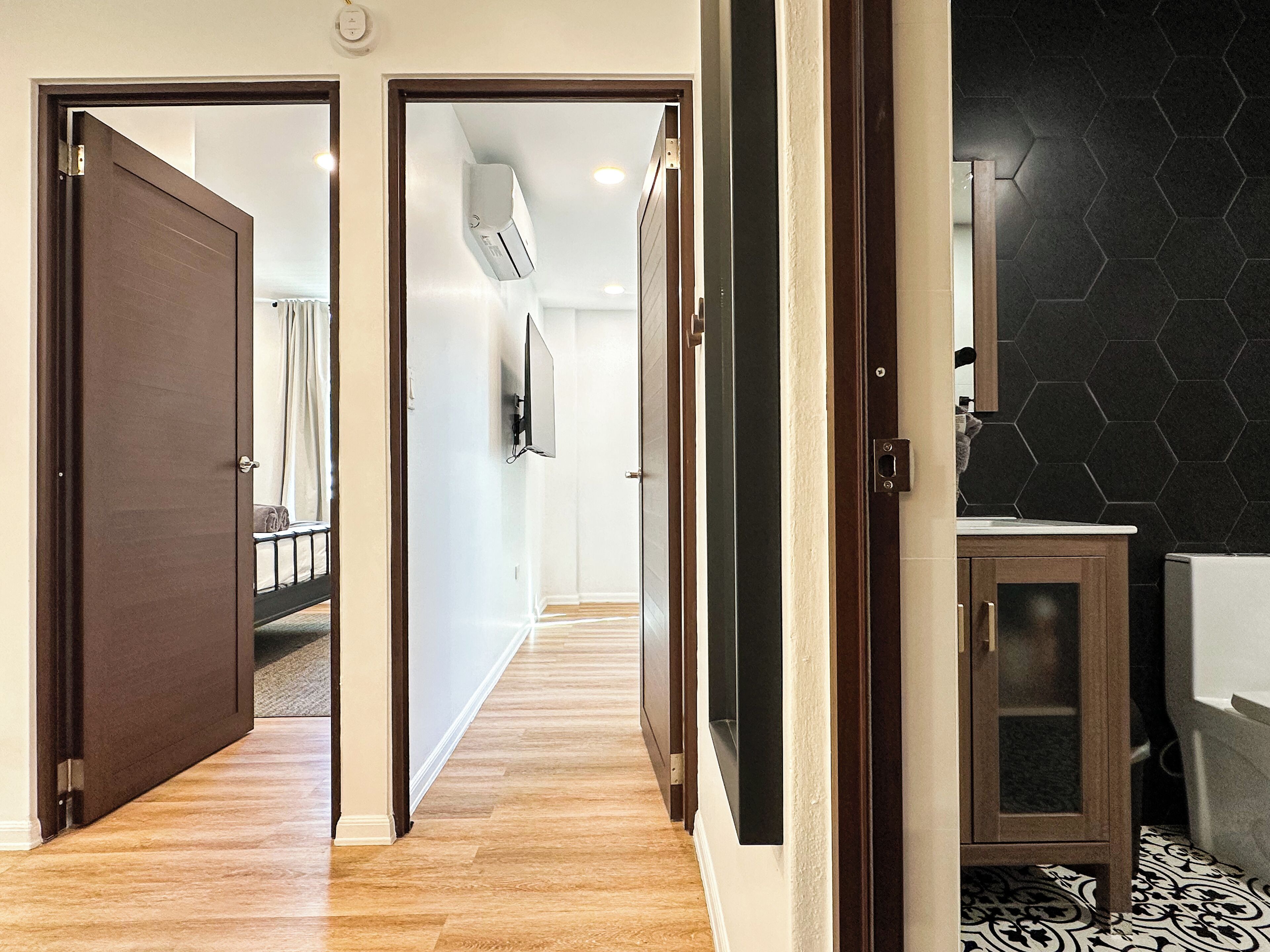 A hallway with wooden flooring, a door leading to a bedroom, and a bathroom with a black hexagonal tile wall.