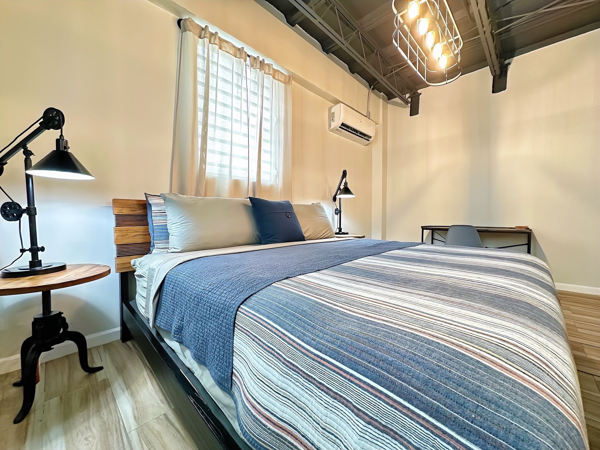 A neatly arranged bedroom with a bed, two bedside tables, a desk, a lamp, a window with curtains, and an air conditioning unit.