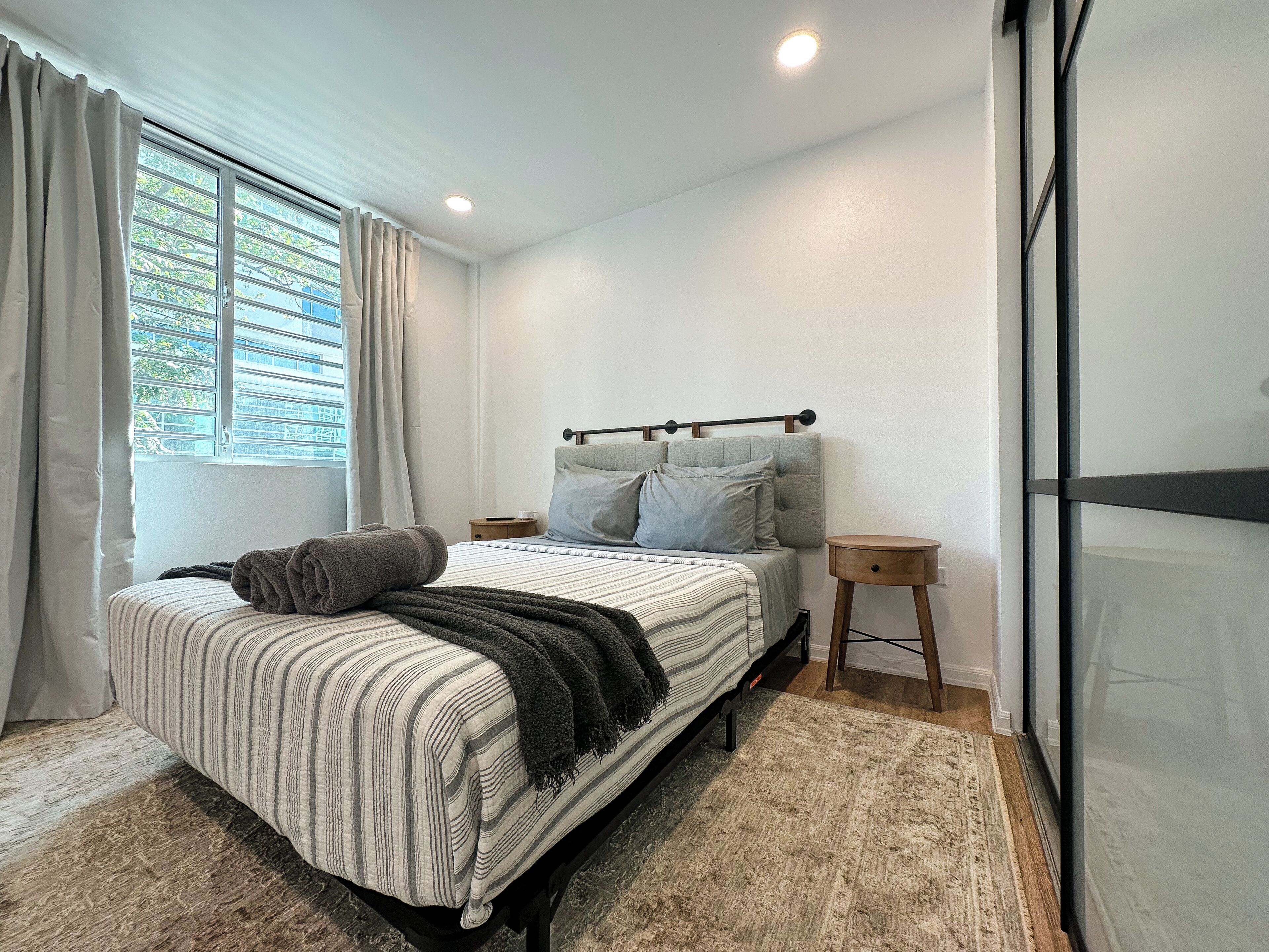 Un dormitorio con cama, mesita de noche y cortinas en la ventana, con vistas a la vegetación.