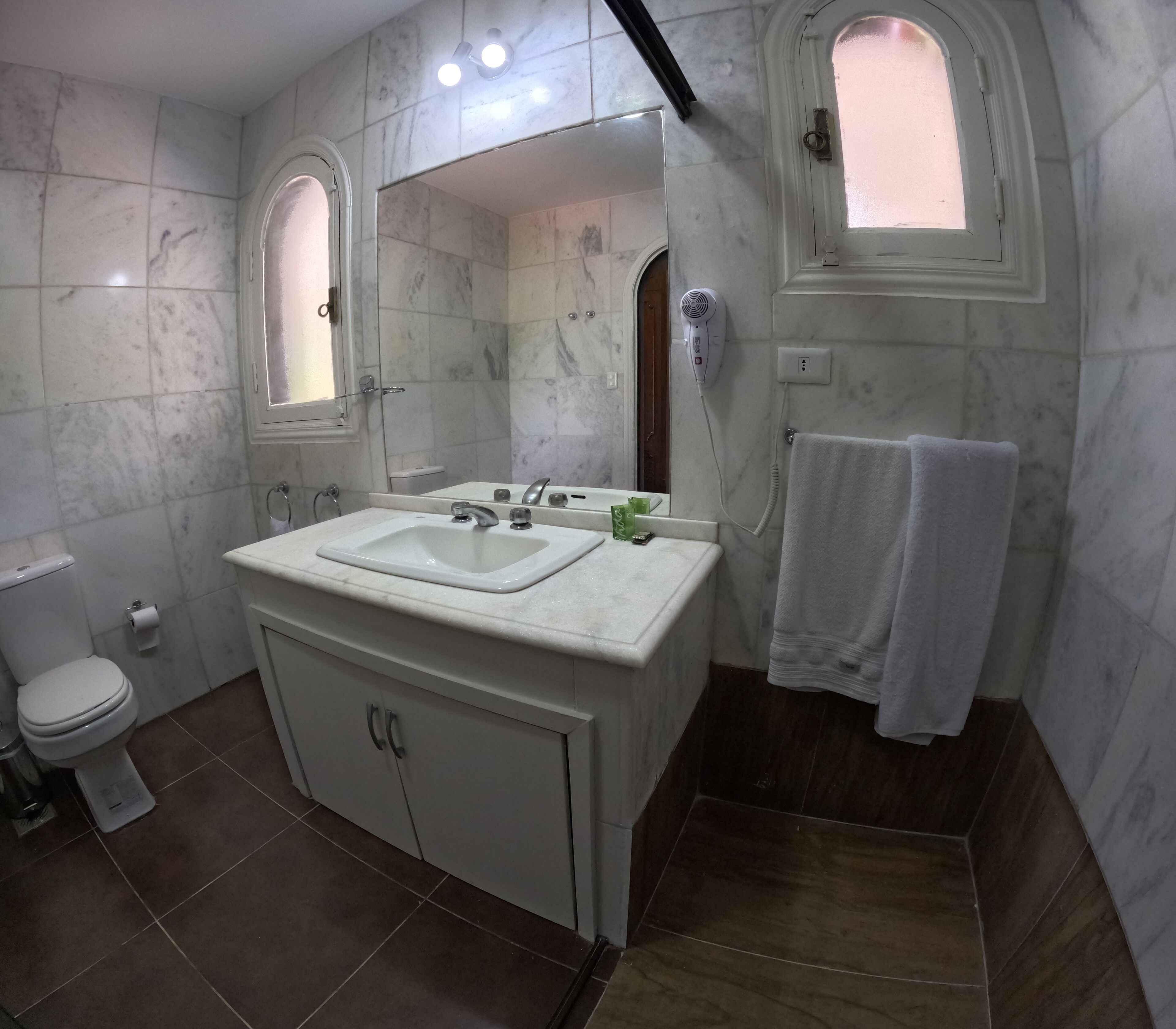 A bathroom with a white sink, mirror, and two vanity mirrors. There is a towel rack with a towel, a toilet, and a hairdryer on the wall.