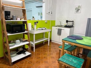 A compact kitchen with a wooden shelf unit, a microwave, a small refrigerator, and a dining table with benches.