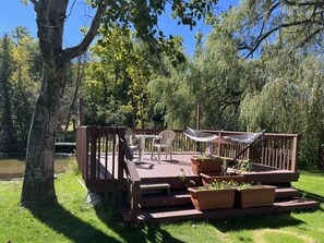 A wooden deck with outdoor furniture, including chairs and a hammock, surrounded by greenery and trees. - Riverview Retreat on the Platte River – Open Year-Round Northern Michigan Magic (Honor)