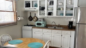 Une cuisine compacte avec des meubles blancs, une cuisinière, un four à micro-ondes et un réfrigérateur. On y trouve également un ventilateur de plafond, un décor mural représentant des poissons et une fenêtre avec des stores.