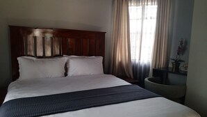 Una cama bien tendida con cabecera de madera, una mesita de noche con lámpara y una ventana con cortinas.