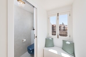 Un baño con inodoro, una ventana con vistas a edificios y un banco con almohadones.