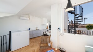 A modern kitchen with white cabinets, a dining area with a wooden table and chairs, and a spiral staircase visible through a window.
