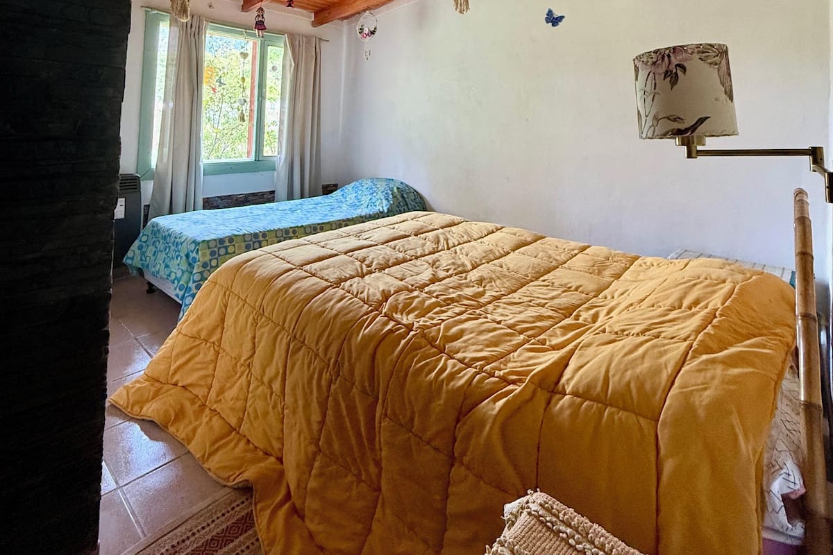 A bedroom with a wooden ceiling, two beds with different bedding, a window with curtains, and a wall-mounted lamp.
