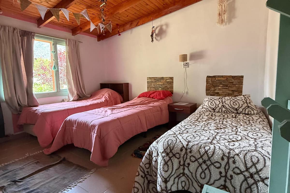 A room with two beds, wooden ceiling beams, a stone wall, and a window with curtains.