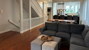 Un salón espacioso con un sofá gris, un reposapiés estampado y una mesa de centro de madera. Una escalera con barandillas blancas conduce a un nivel superior.