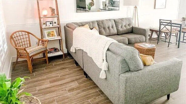 A living room with a grey sofa, a wicker chair, a bookshelf, and a 'Beach' sign on the wall.