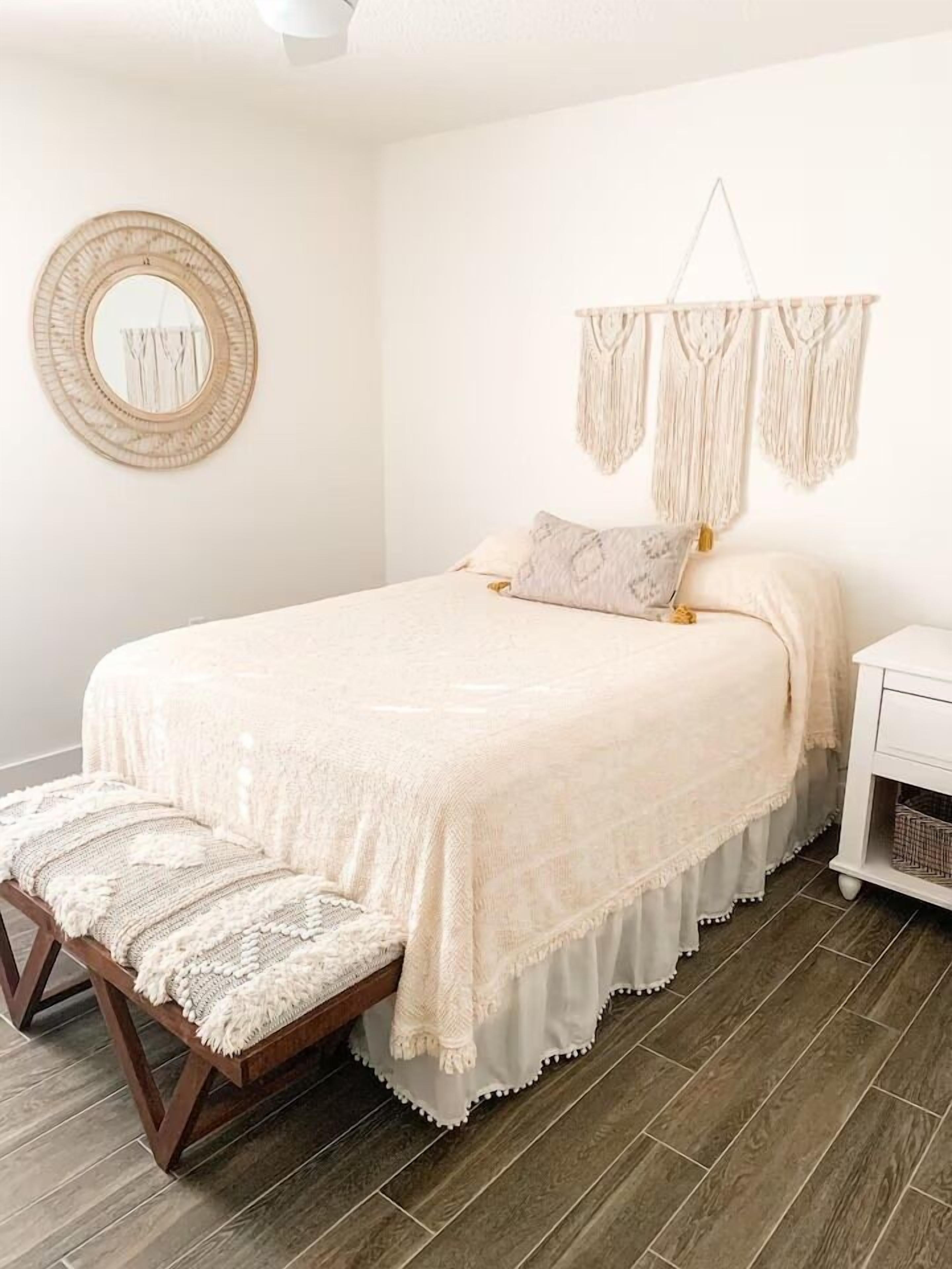 A neatly made bed with a white bedspread and pillows, a wooden bench with a white cushion, a round mirror, and macramé wall hangings.