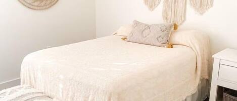 A neatly made bed with a white bedspread and pillows, a wooden bench with a white cushion, a round mirror, and macramé wall hangings.