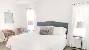 A large bed with a dark headboard, two patterned pillows, and a light-colored bedspread. There are two wicker chairs and a small table with a lamp.