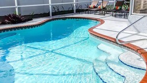 A swimming pool with a brick border, surrounded by lounge chairs and a covered area with a blue umbrella.