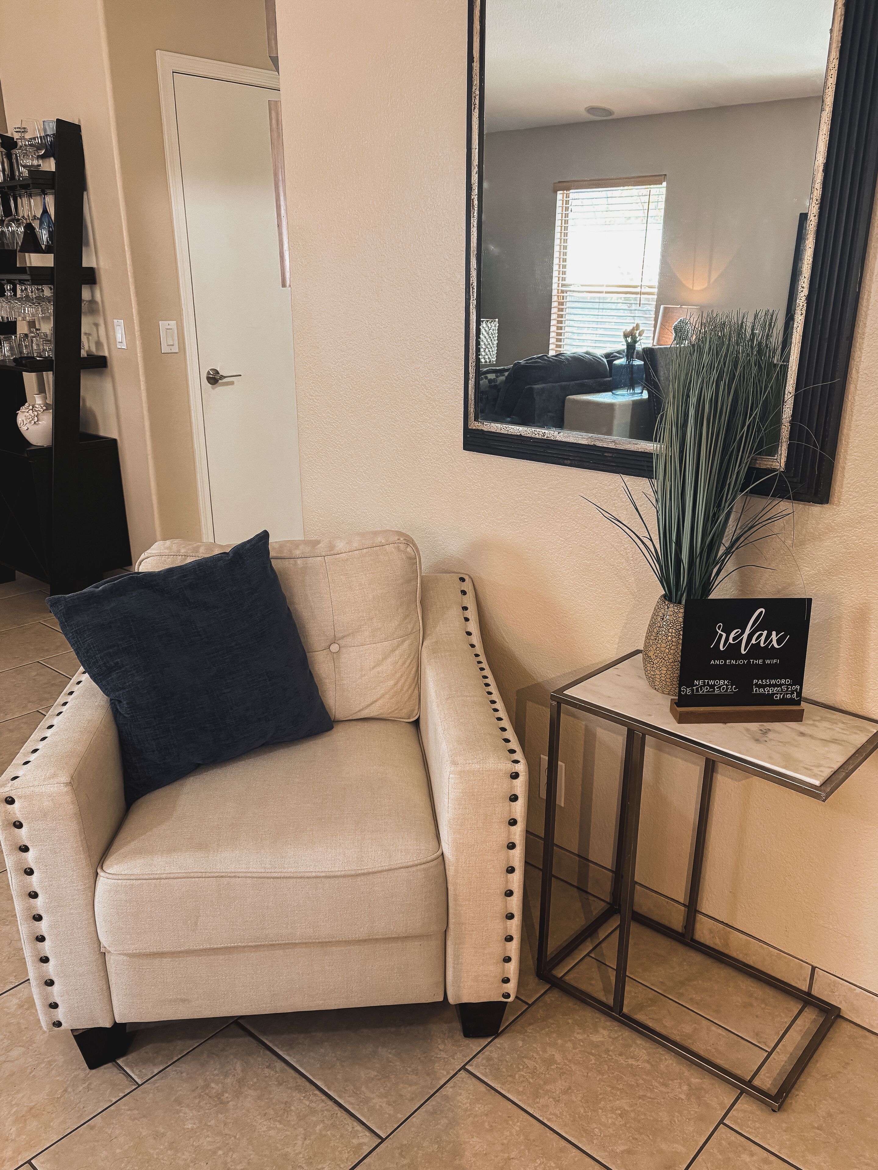 Un fauteuil beige avec un coussin bleu foncé, une petite table d’appoint sur laquelle est posé un panneau, et une grande plante verte.